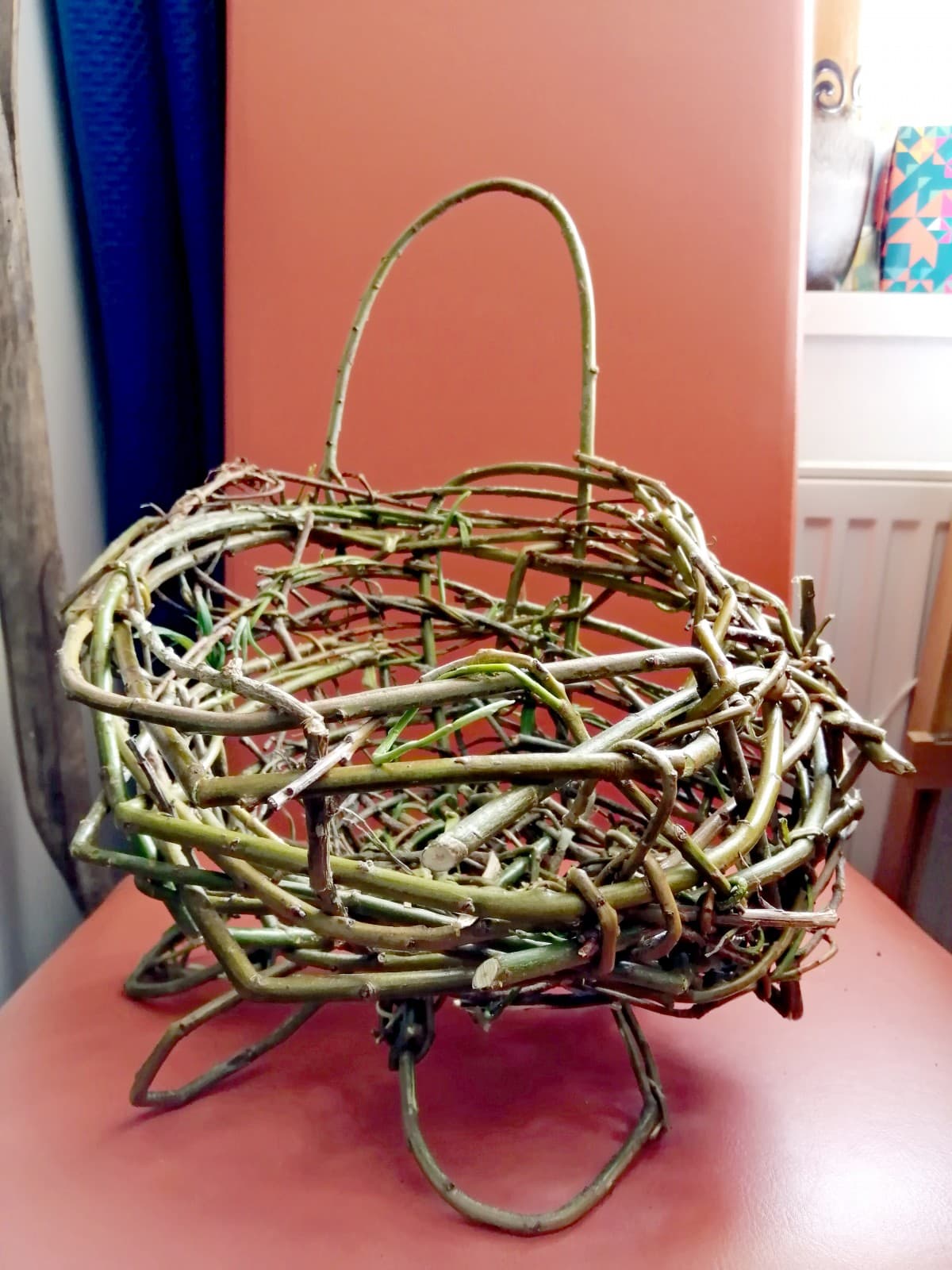 The bare bones of the willow basket sitting on an orange chair. It looks quite like a birds nest!